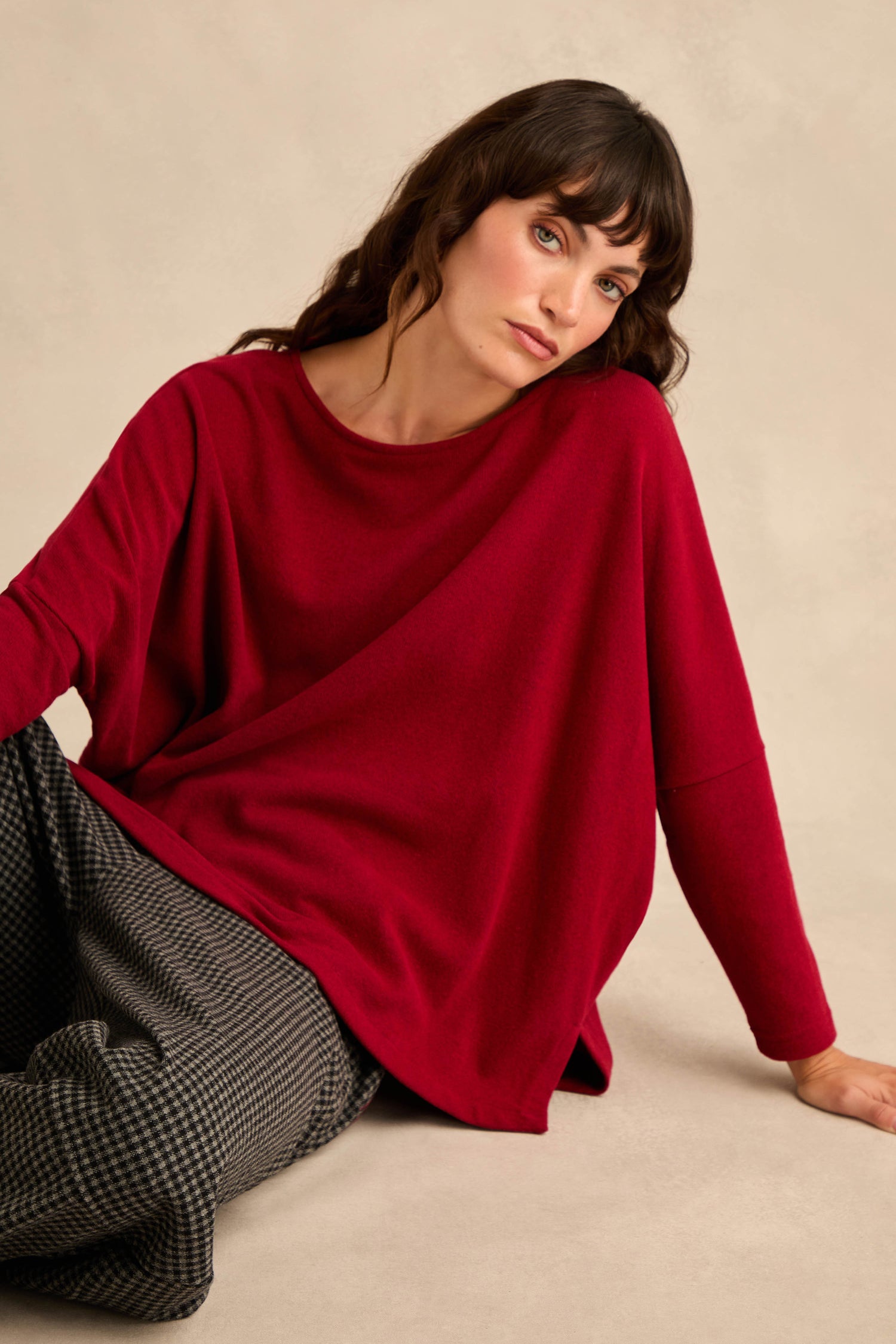 A woman with wavy brown hair sits on the floor against a beige background, wearing the Round Neck Soft Jersey Top in red and checkered gray pants.