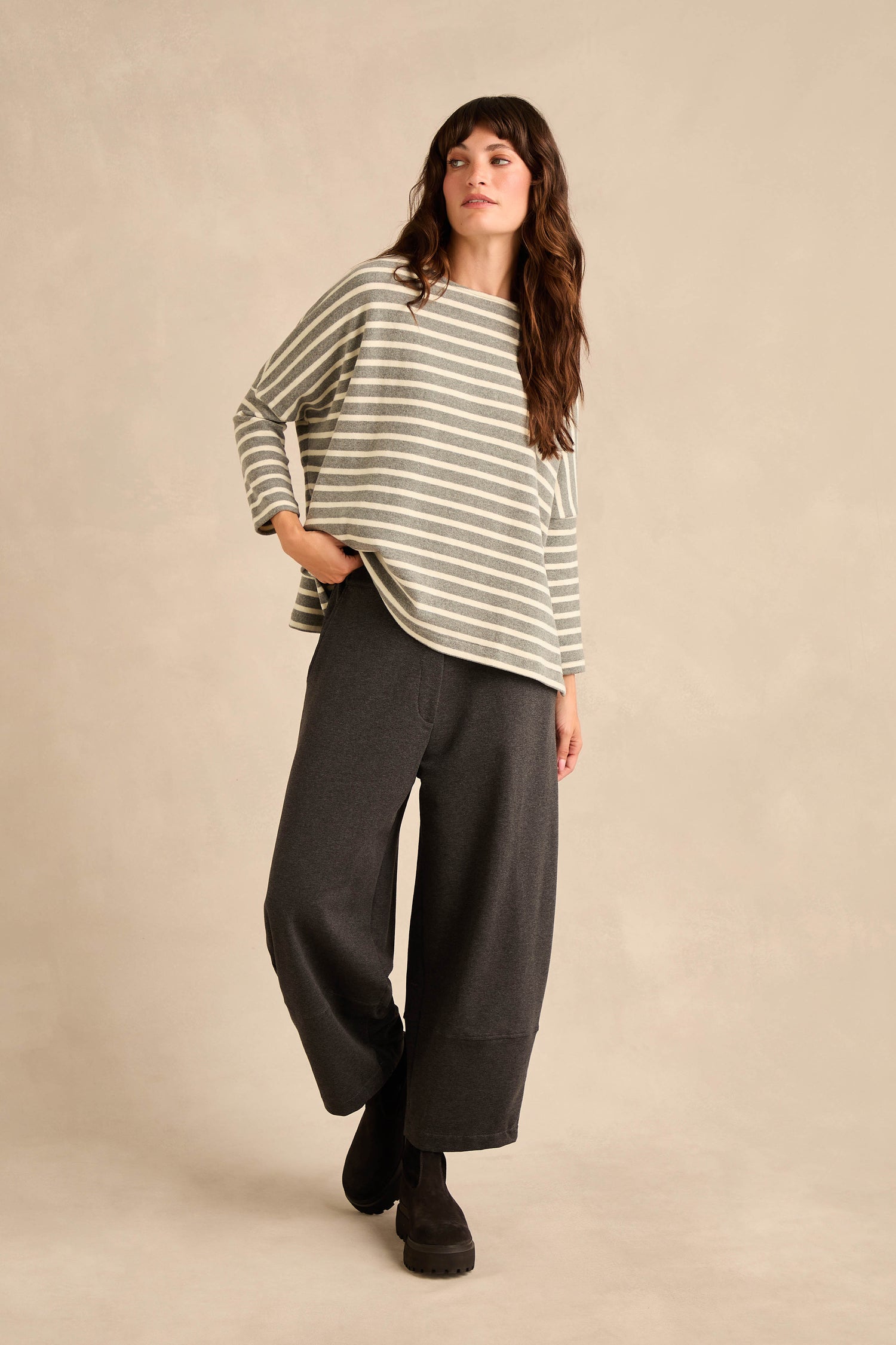 A woman stands against a plain beige background wearing the Round Neck Wide Stripe Jersey Top, made from a cotton-blend jersey, paired with wide-leg dark pants.