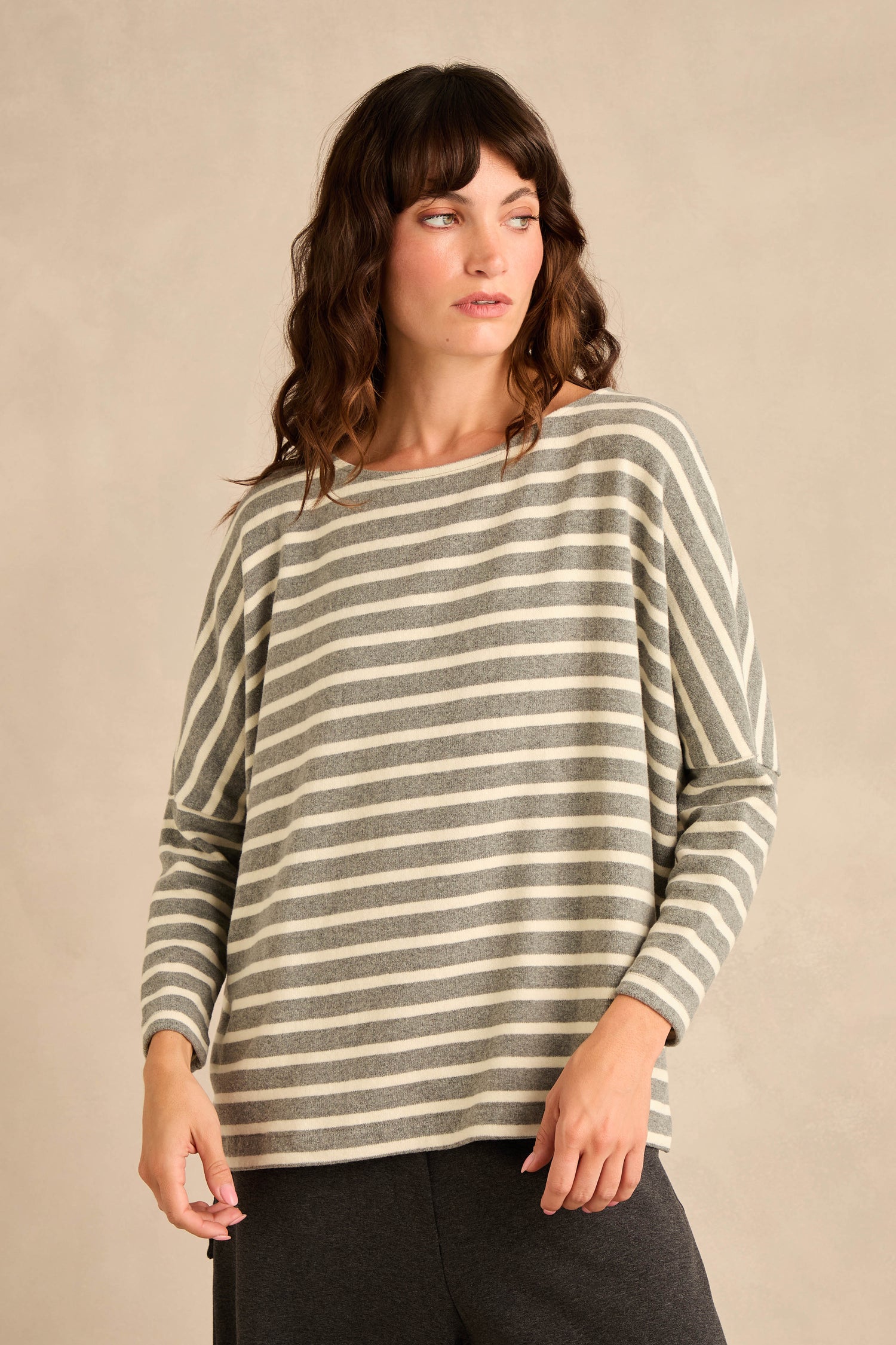 A woman stands against a plain beige background, looking to her left, wearing the Round Neck Wide Stripe Jersey Top—a long-sleeved jersey in soft cotton-blend with classic gray and white wide stripes and a round neckline—paired with dark pants.
