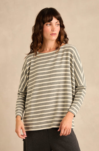 A woman stands against a plain beige background, looking to her left, wearing the Round Neck Wide Stripe Jersey Top—a long-sleeved jersey in soft cotton-blend with classic gray and white wide stripes and a round neckline—paired with dark pants.