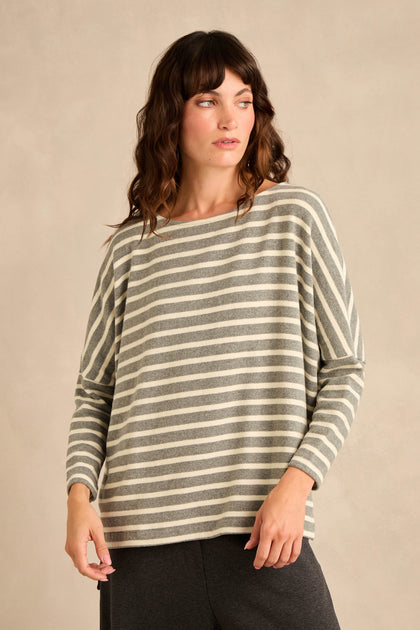A woman stands against a plain beige background, looking to her left, wearing the Round Neck Wide Stripe Jersey Top—a long-sleeved jersey in soft cotton-blend with classic gray and white wide stripes and a round neckline—paired with dark pants.