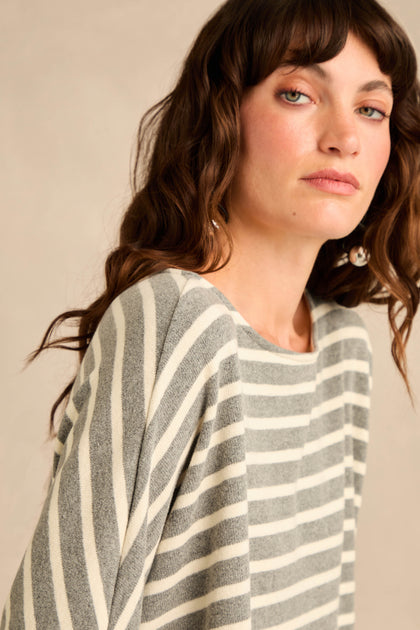 A woman with wavy brown hair, gazing slightly to the side against a neutral background, is wearing the Round Neck Wide Stripe Jersey Top. The gray and white stripes and soft cotton-blend jersey fabric lend her outfit a relaxed, comfortable feel.