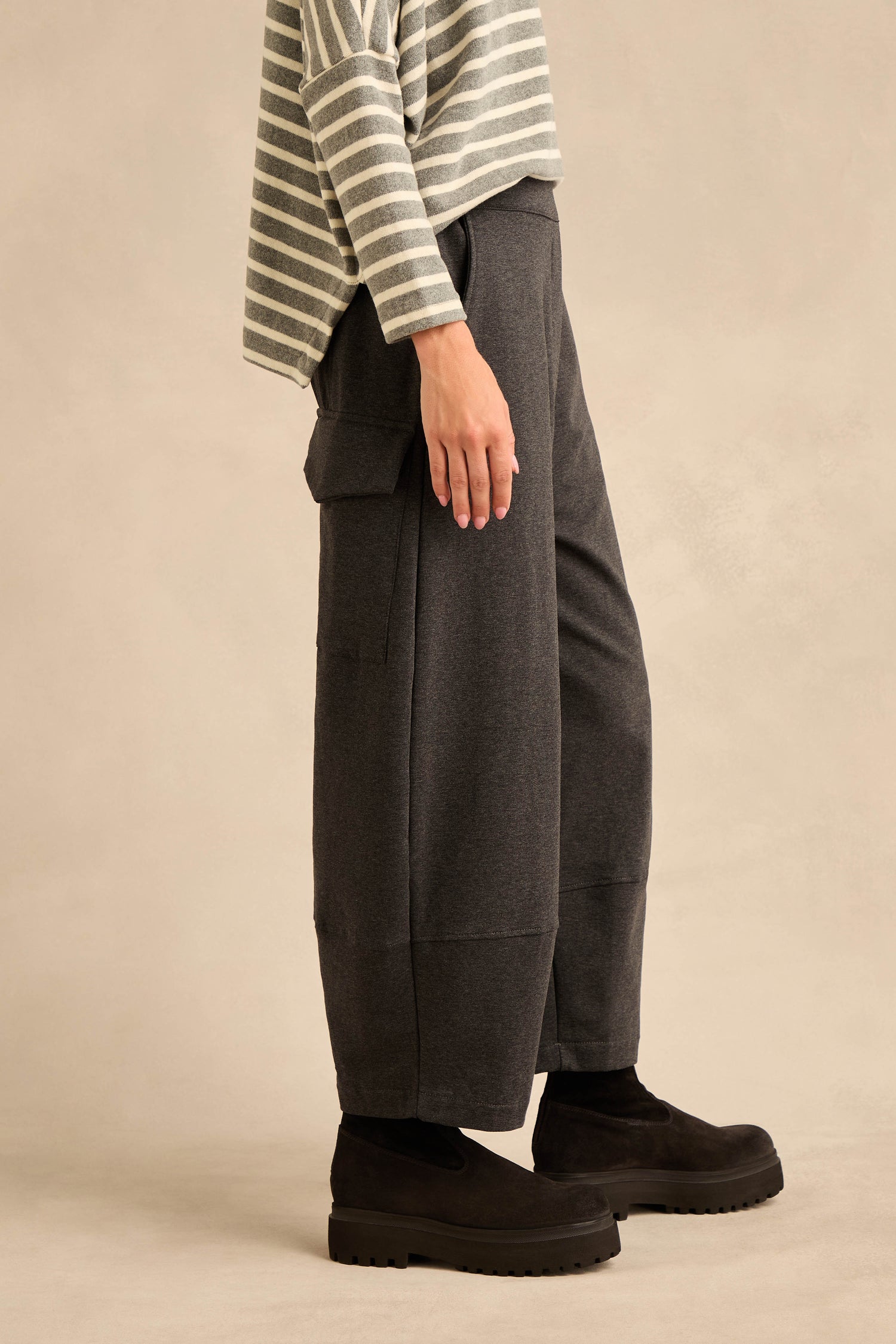 A person stands sideways against a plain beige background, wearing a gray and white striped long-sleeve top, Jersey Pocket Trousers in dark gray cotton stretch jersey with pockets, and black platform shoes.
