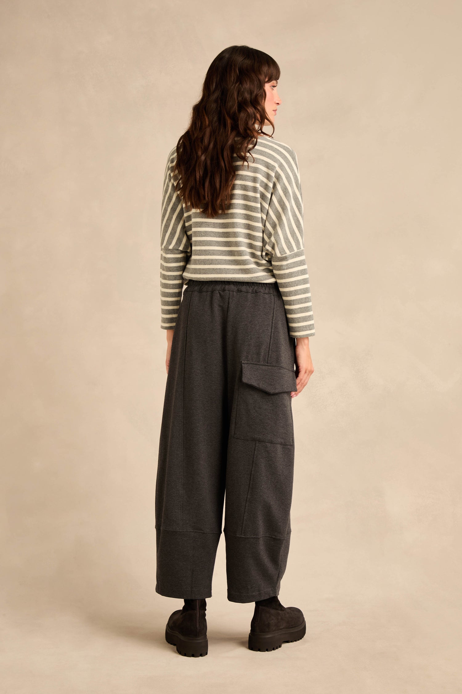 A person stands facing away, wearing a striped long-sleeve top paired with the Jersey Pocket Trousers—relaxed, wide dark gray trousers featuring a side pocket—and black shoes against a neutral background.