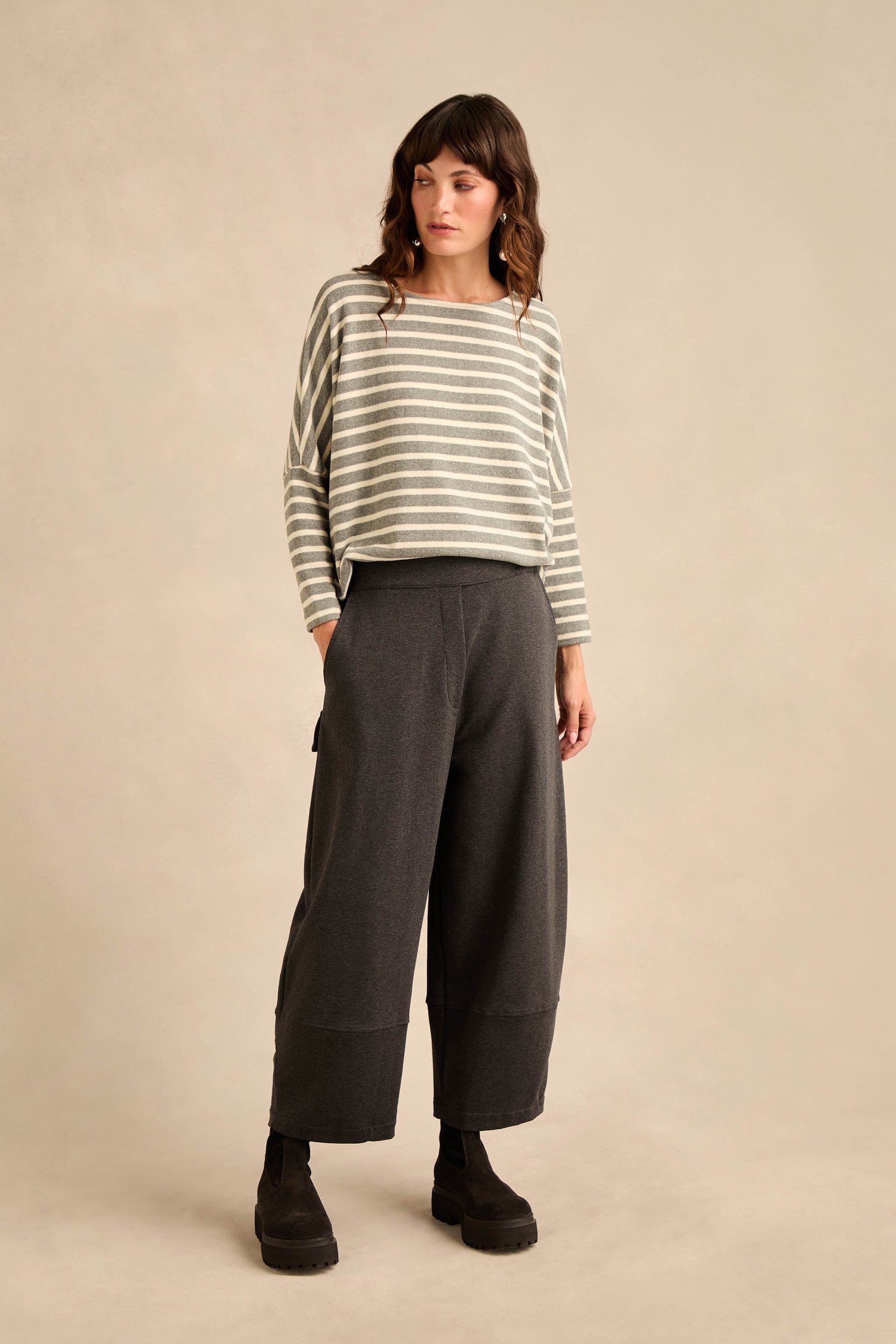 A person is wearing a light gray and white striped long-sleeve shirt, black boots, and dark Jersey Pocket Trousers made from relaxed cotton stretch jersey, standing against a plain beige background.