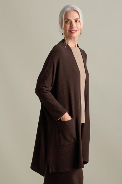 A woman with gray hair is standing, wearing a Souffle Soft Jersey Cardi in longline length brown over a beige top. She has her hands in the cardigan pockets and is looking off to the side while smiling.