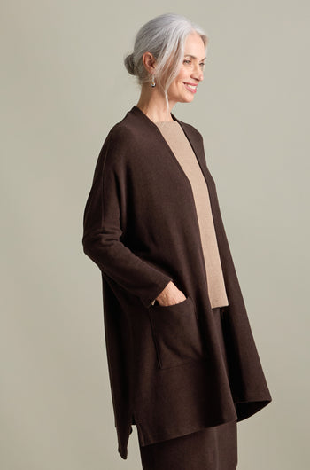 A woman with gray hair is wearing a Souffle Soft Jersey Cardi in a longline length over a beige top, standing against a neutral background. She smiles slightly with her left hand in her cardigan pocket.