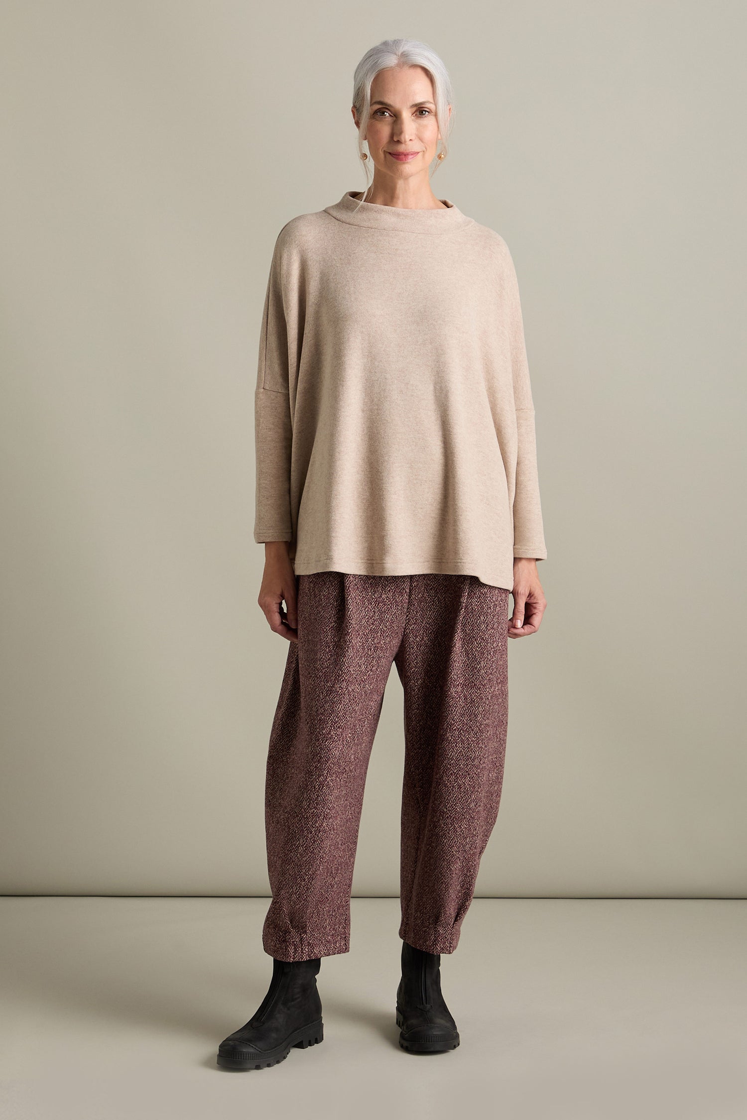 A woman with gray hair stands against a neutral background, wearing an Italian-crafted Spino Boxy Soft Jersey Top in beige, maroon textured pants, and black boots.