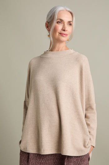 A woman with gray hair, dressed in a beige Spino Boxy Soft Jersey Top from Mama B and patterned pants, stands against a neutral background. She is smiling slightly and looking to the side.