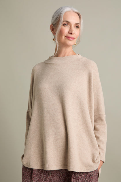 A woman with gray hair, dressed in a beige Spino Boxy Soft Jersey Top from Mama B and patterned pants, stands against a neutral background. She is smiling slightly and looking to the side.