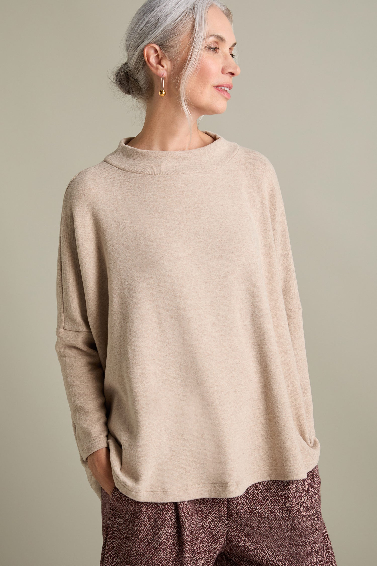 A person wearing a beige Mama B Spino Boxy Soft Jersey Top, crafted in Italy, along with maroon pants is looking to the side. Their hair is tied back, and they are adorned with gold earrings. The background is plain and neutral.