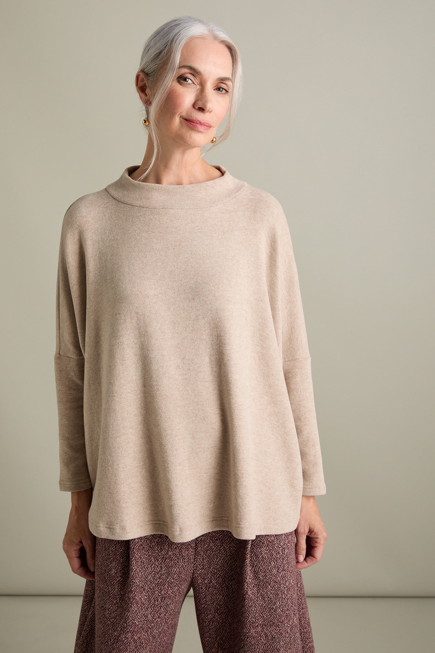 A woman with grey hair, wearing a beige oversized top from the Spino Boxy Soft Jersey Top collection and brown patterned pants, stands against a plain background.