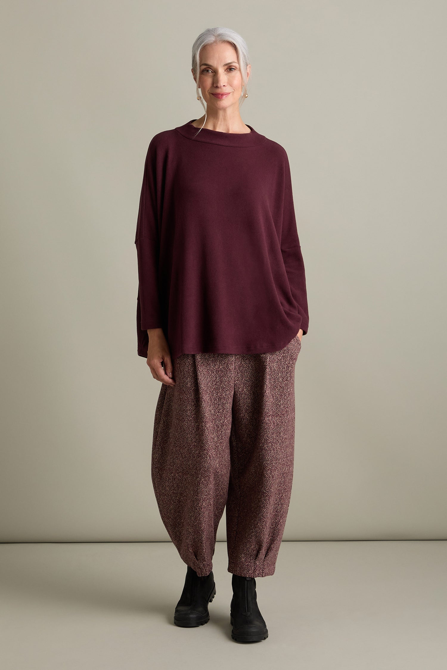 A woman with gray hair stands against a neutral background, wearing an Italian-crafted, oversized Spino Boxy Soft Jersey Top in maroon by Mama B, patterned baggy pants, and black boots. She smiles slightly, with one hand in her pocket.