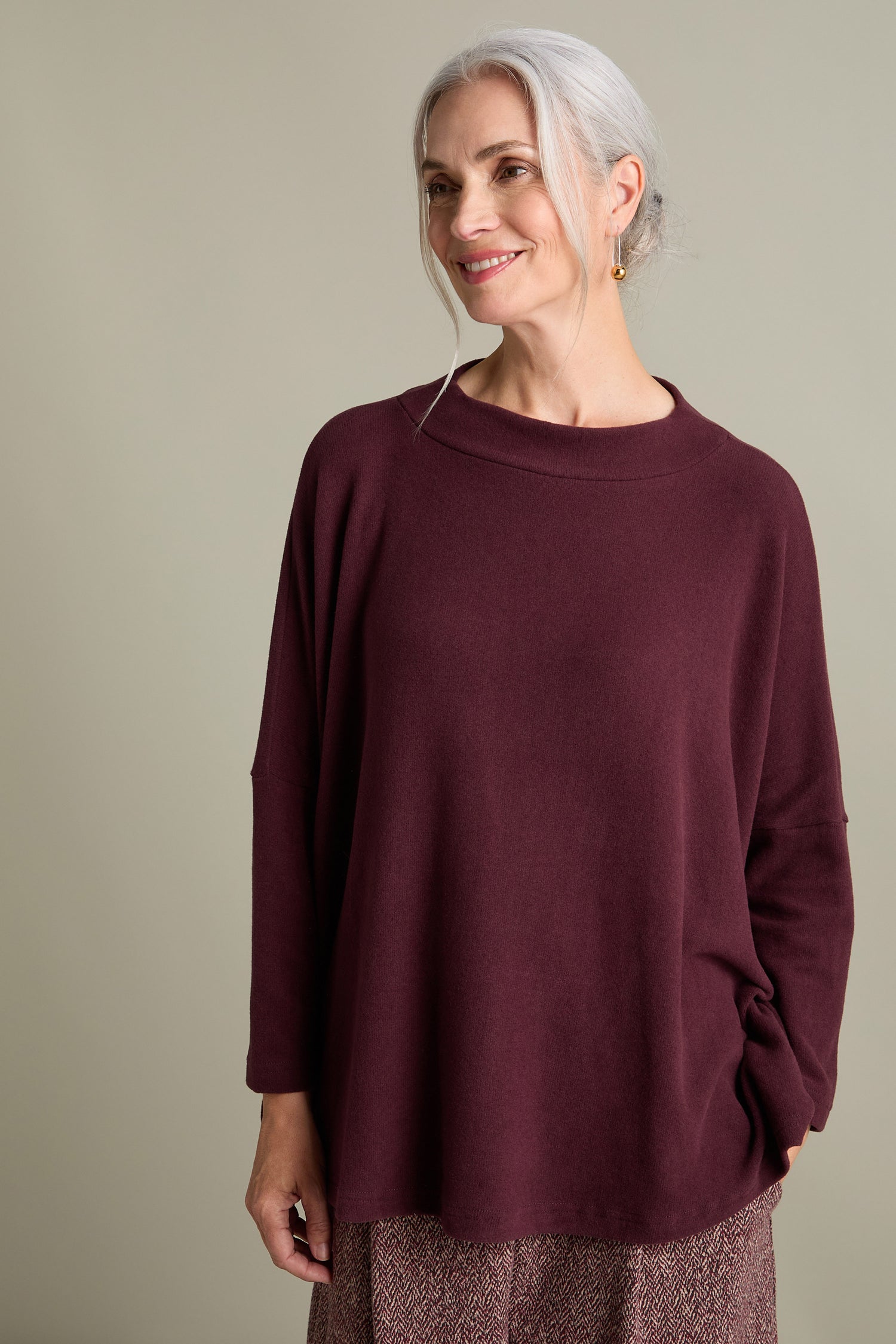 A person with gray hair and a smile poses in the Italian-crafted Spino Boxy Soft Jersey Top and patterned pants against a beige background.