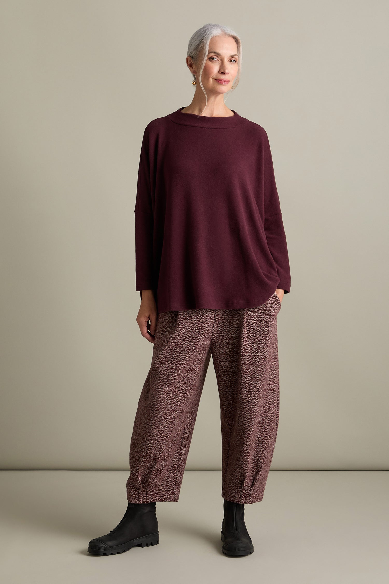 An older woman with gray hair stands in a relaxed pose, wearing an Italian-crafted Spino Boxy Soft Jersey Top in burgundy, patterned loose pants, and black boots. The background is a plain light color.