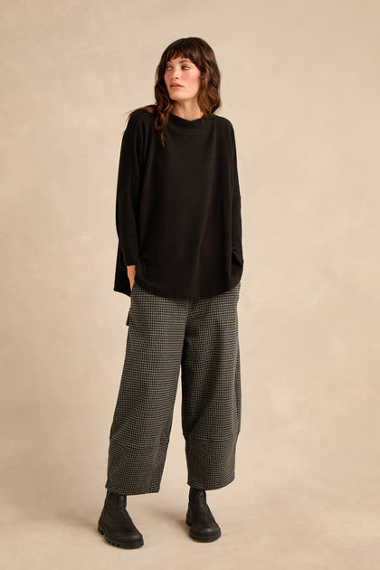 A person stands against a beige background wearing the Black Soft Jersey Boxy Top with a boxy silhouette, paired with wide checkered pants and black boots, hands in pockets and looking to the side.