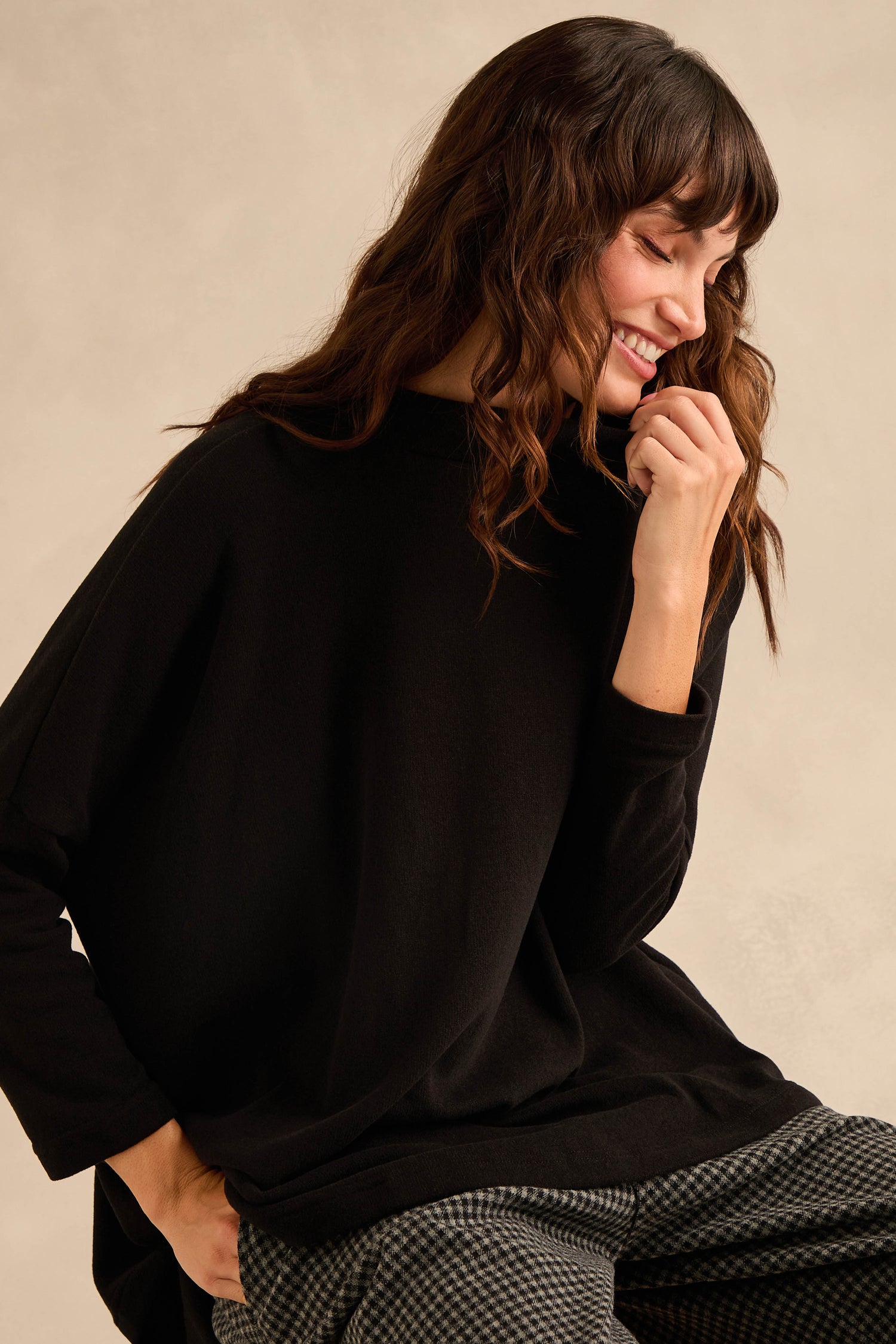 A woman with long wavy brown hair wears the Black Soft Jersey Boxy Top, featuring a boxy silhouette, paired with checked pants. She smiles and looks down with one hand near her face against a neutral background.