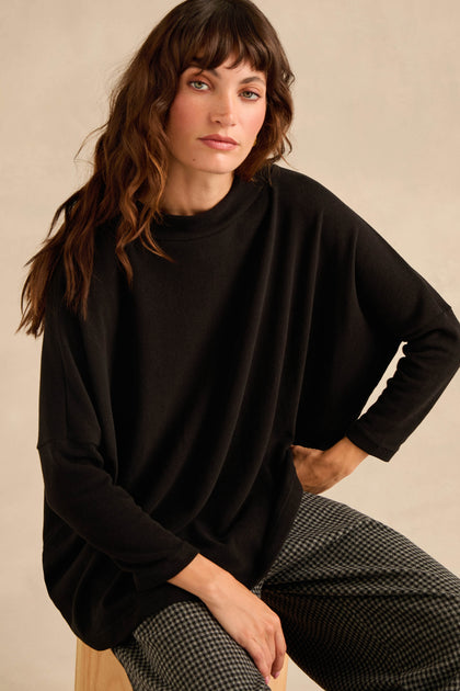 A woman with wavy brown hair sits on a stool against a neutral background, wearing the Black Soft Jersey Boxy Top in a loose, boxy silhouette paired with checkered pants.