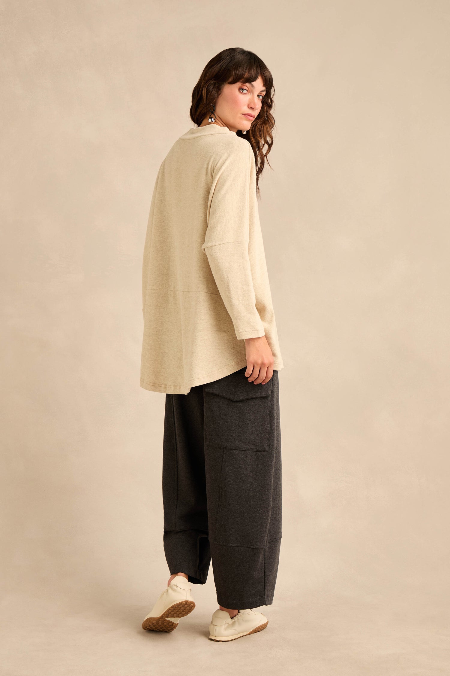 Person standing on a neutral background, wearing the Cream Marl Soft Jersey Boxy Top with a relaxed fit, paired with dark wide-leg pants with large pockets and white sneakers, facing slightly away from the camera.