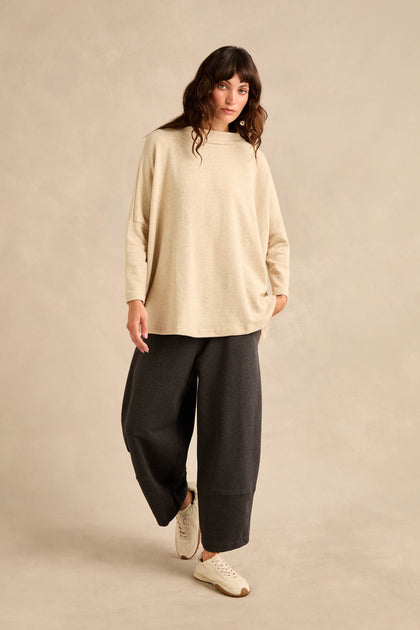 A person stands against a beige background wearing the Cream Marl Soft Jersey Boxy Top in a relaxed fit, paired with wide dark pants and white sneakers, with one hand in their pocket.