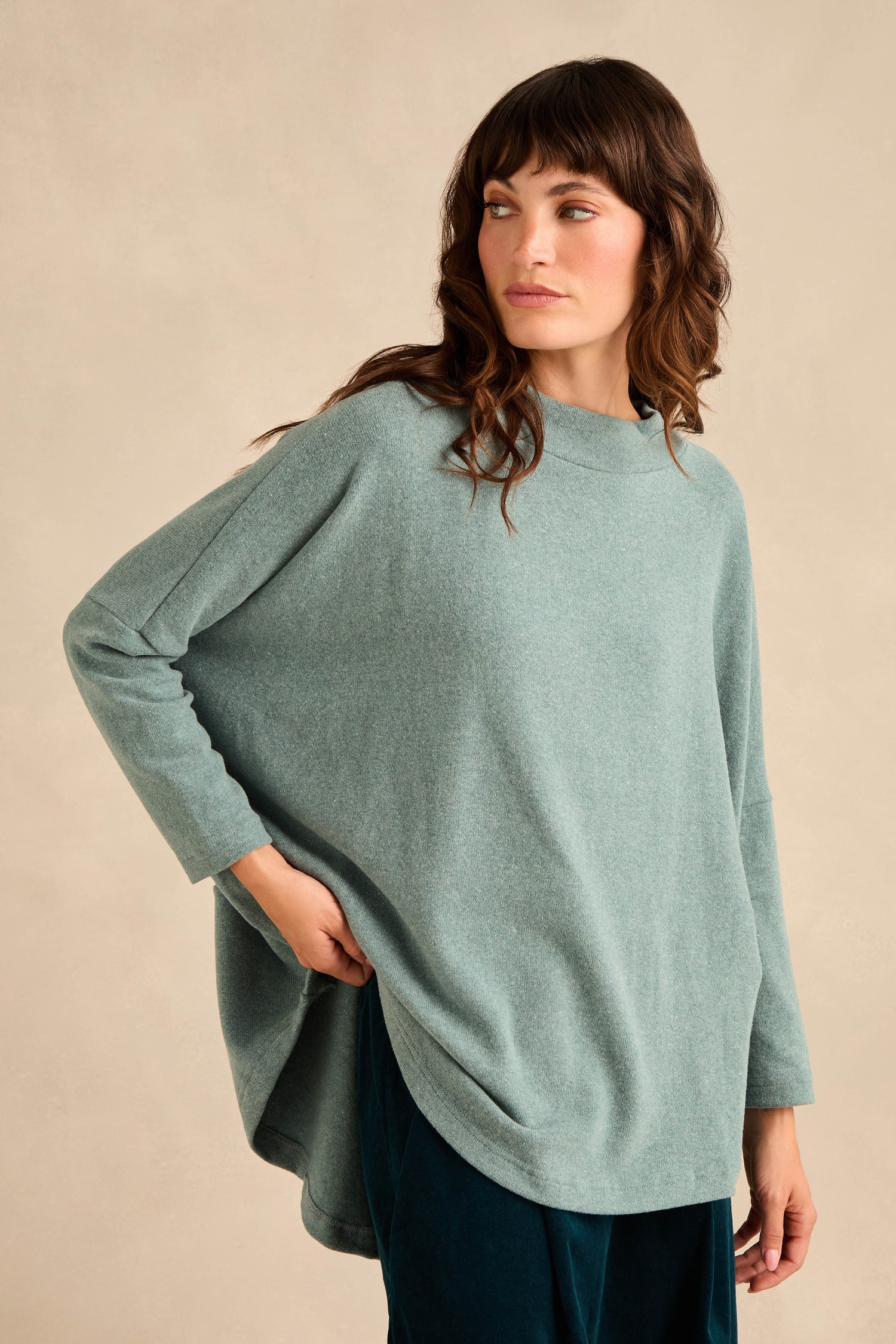 A woman with wavy brown hair, wearing the Teal Soft Jersey Boxy Top and dark pants, stands against a beige background looking to the side.
