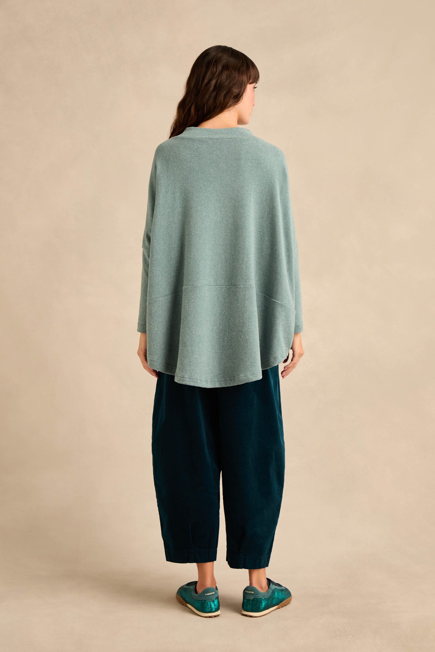 A woman stands with her back to the camera, wearing the Teal Soft Jersey Boxy Top—a loose, cotton-blend light blue sweater with a boxy silhouette—paired with wide dark teal pants and teal shoes against a plain beige background.