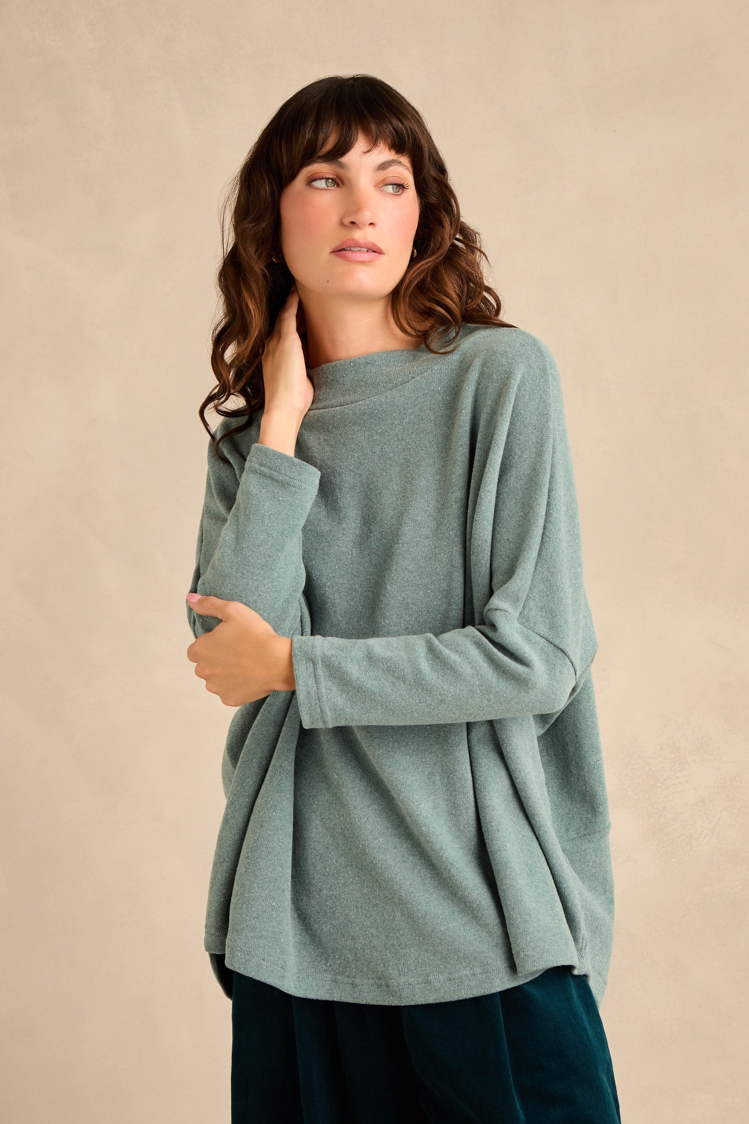A woman wearing the Teal Soft Jersey Boxy Top, featuring a loose fit, long sleeves, and a boxy silhouette, stands against a neutral background, gazing to her left with one hand touching her neck.