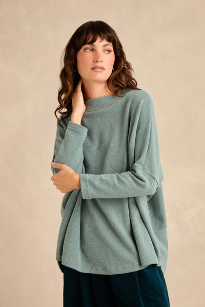 A woman wearing the Teal Soft Jersey Boxy Top, featuring a loose fit, long sleeves, and a boxy silhouette, stands against a neutral background, gazing to her left with one hand touching her neck.
