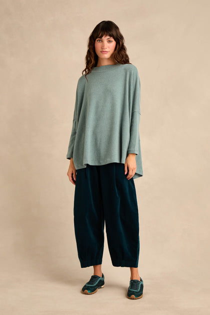A woman with wavy brown hair and a neutral expression stands against a beige background, wearing the Teal Soft Jersey Boxy Top—a loose, cotton-blend light green long-sleeve top with a boxy silhouette—paired with wide dark green pants and teal sneakers.