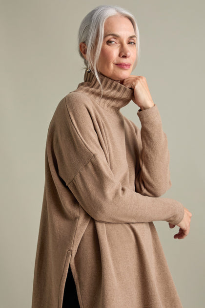 An older woman with gray hair, wearing a taupe Zeppola Rib Neck Soft Jersey Tunic, stands with her arm bent and hand touching her chin, against a beige background.