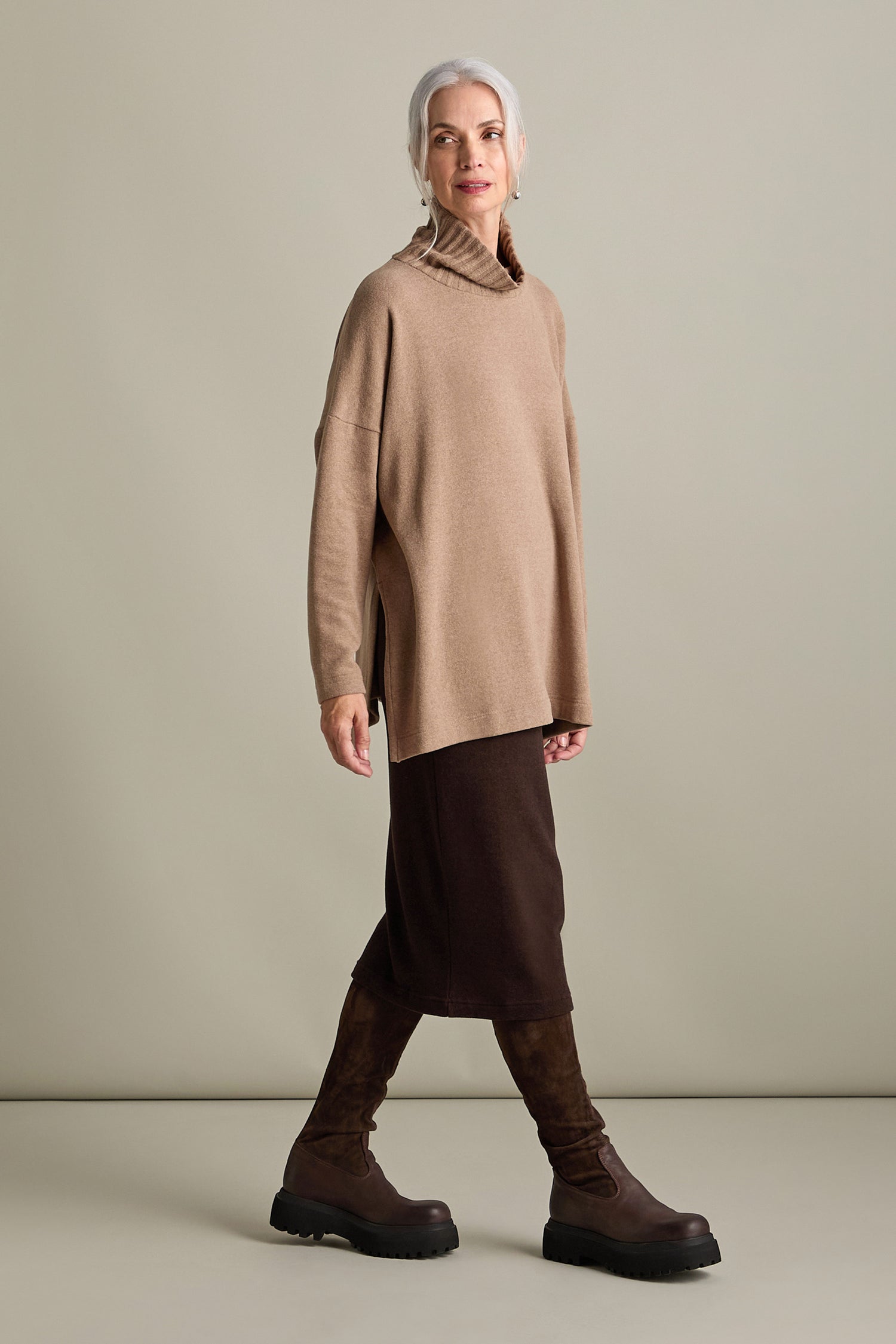 A woman with grey hair is wearing an oversized Zeppola Rib Neck Soft Jersey Tunic in brown, complemented by a dark brown skirt and brown knee-high boots, standing against a plain background.