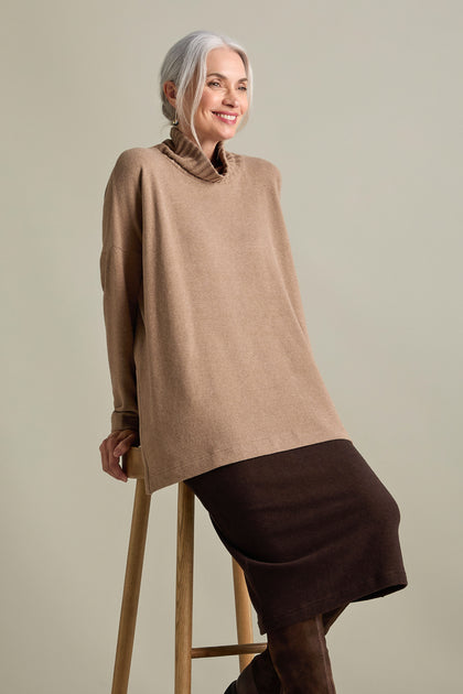 A person with gray hair, wearing a beige Zeppola Rib Neck Soft Jersey Tunic and a brown skirt, sits on a wooden stool against a plain background.