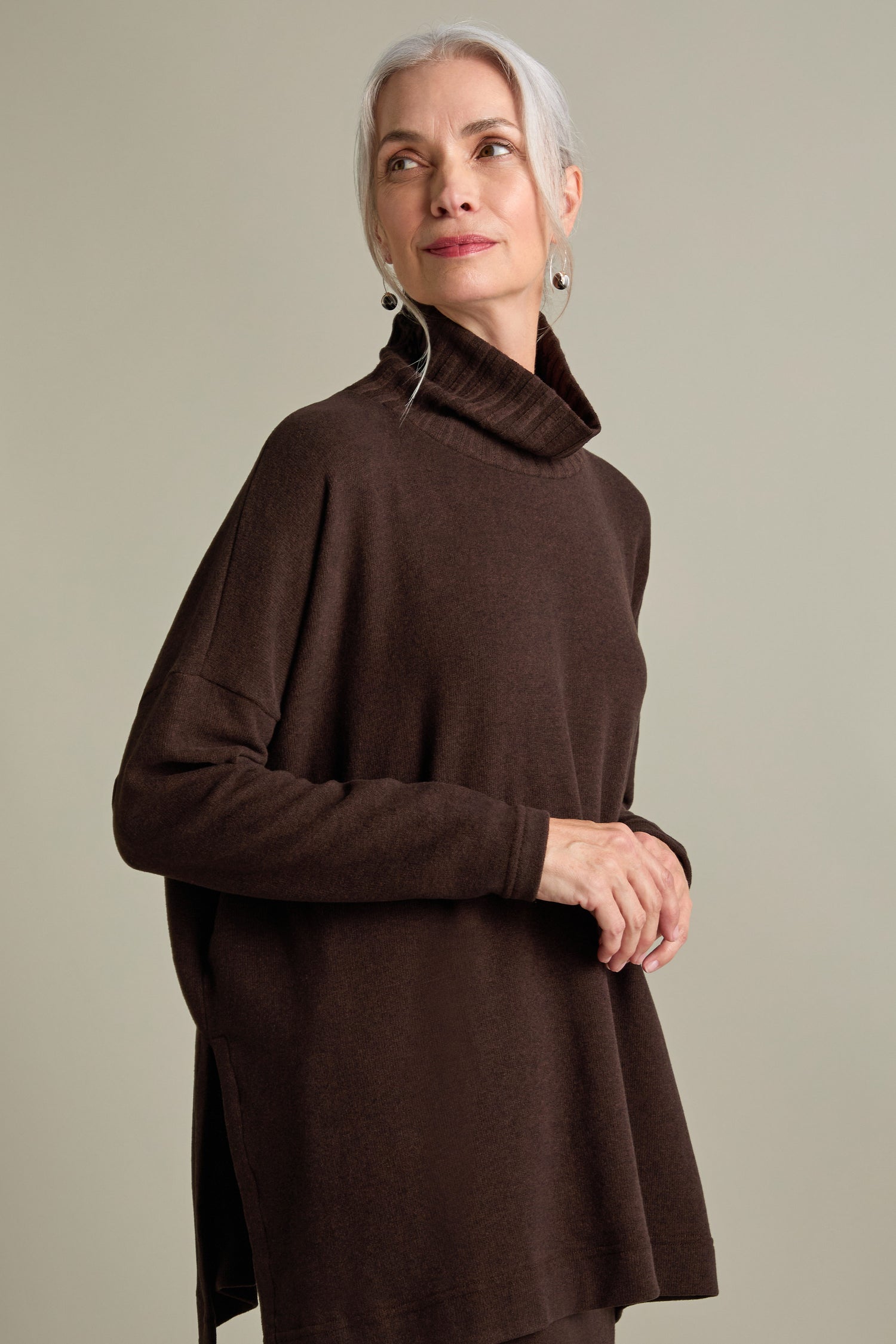 A woman with gray hair in a bun is wearing a brown Zeppola Rib Neck Soft Jersey Tunic. She is standing against a plain background, looking upwards to her right with a neutral expression.