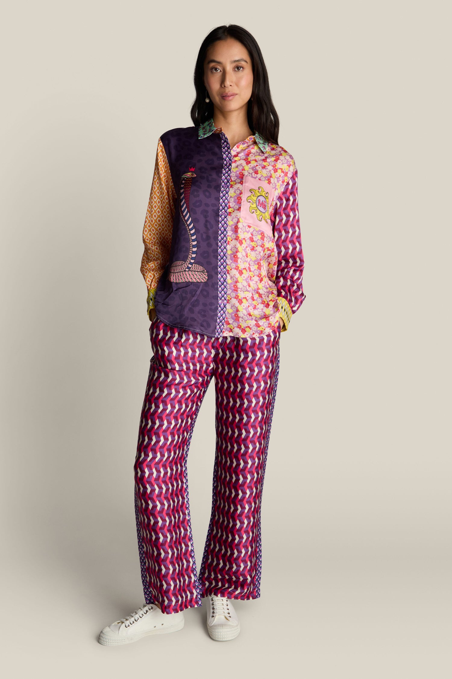 A woman stands against a neutral background wearing a colorful, bold print outfit that includes a long-sleeve shirt and matching Twisted Love Bailey Trousers, paired with white sneakers.