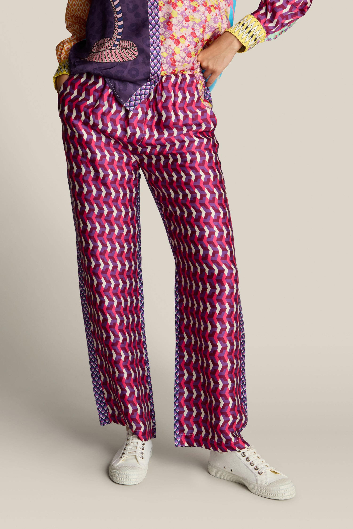 Person wearing a multi-colored bold print design shirt and Twisted Love Bailey Trouser in a pink geometric pattern, paired with white sneakers, standing with one hand on their hip against a plain background.