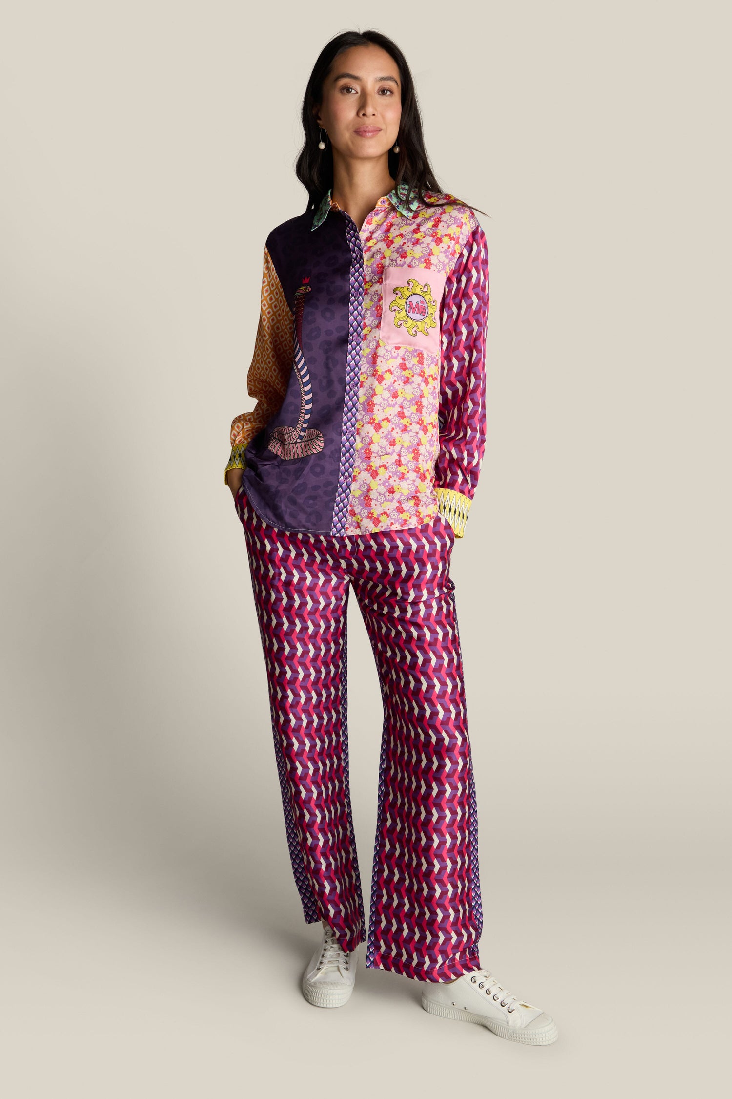 A woman stands against a plain background wearing a colorful, bold print design shirt and matching Twisted Love Bailey Trousers paired with white sneakers. She has her hands in her pockets and is looking slightly off-camera.