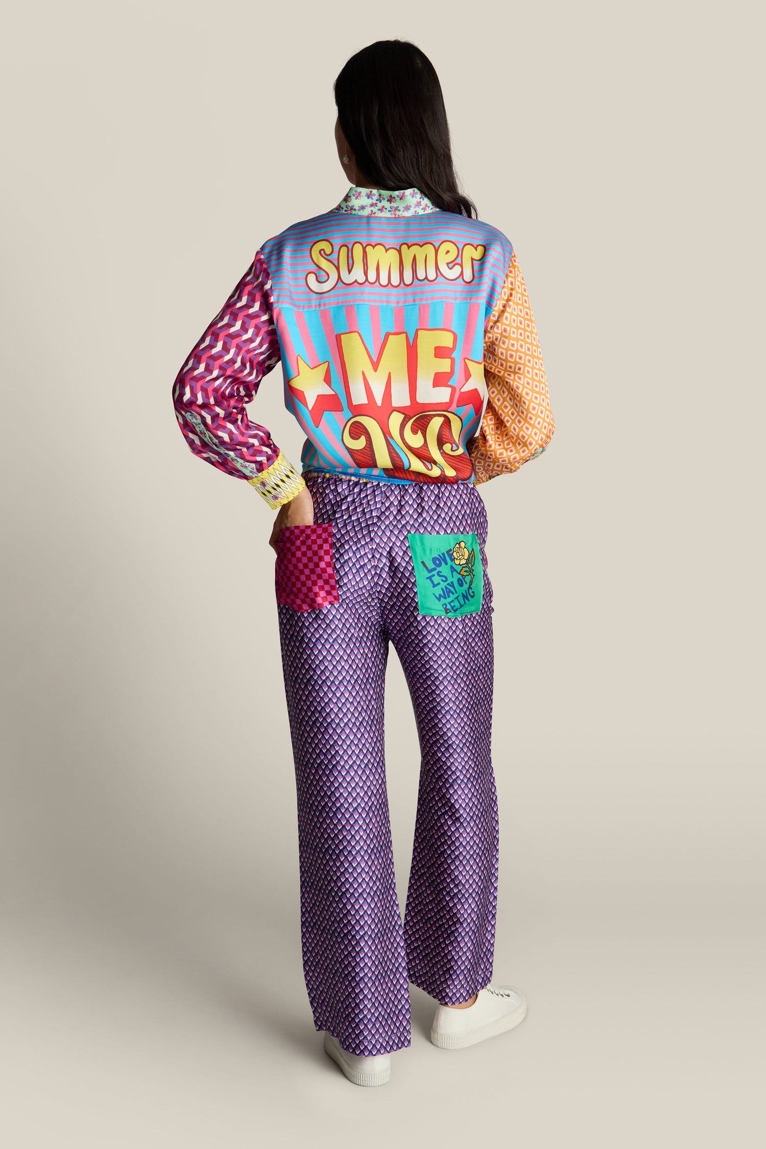 A person with dark hair stands facing away from the camera, wearing a vibrant outfit with "Summer Me" emblazoned on the back of their jacket. The bold print design catches the eye as they flaunt a pair of Twisted Love Bailey Trousers in a straight-leg silhouette. They complete their look with white shoes.