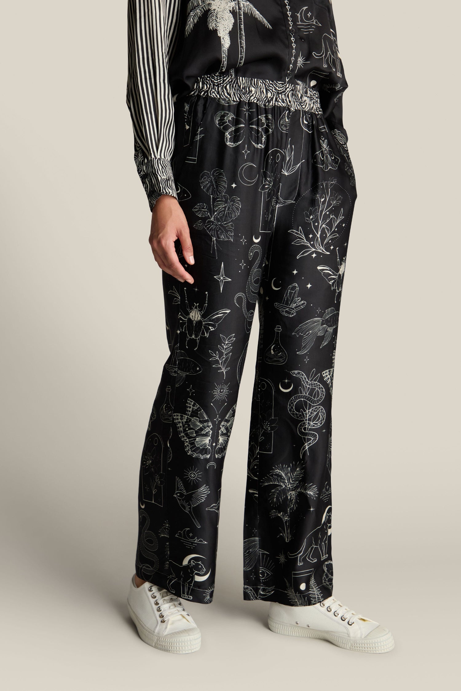 A person wearing a black outfit with botanical and celestial prints and white sneakers is standing upright. Only the lower half of the body, featuring Black Magic Bailey Trousers with an elasticated waistband, is visible.