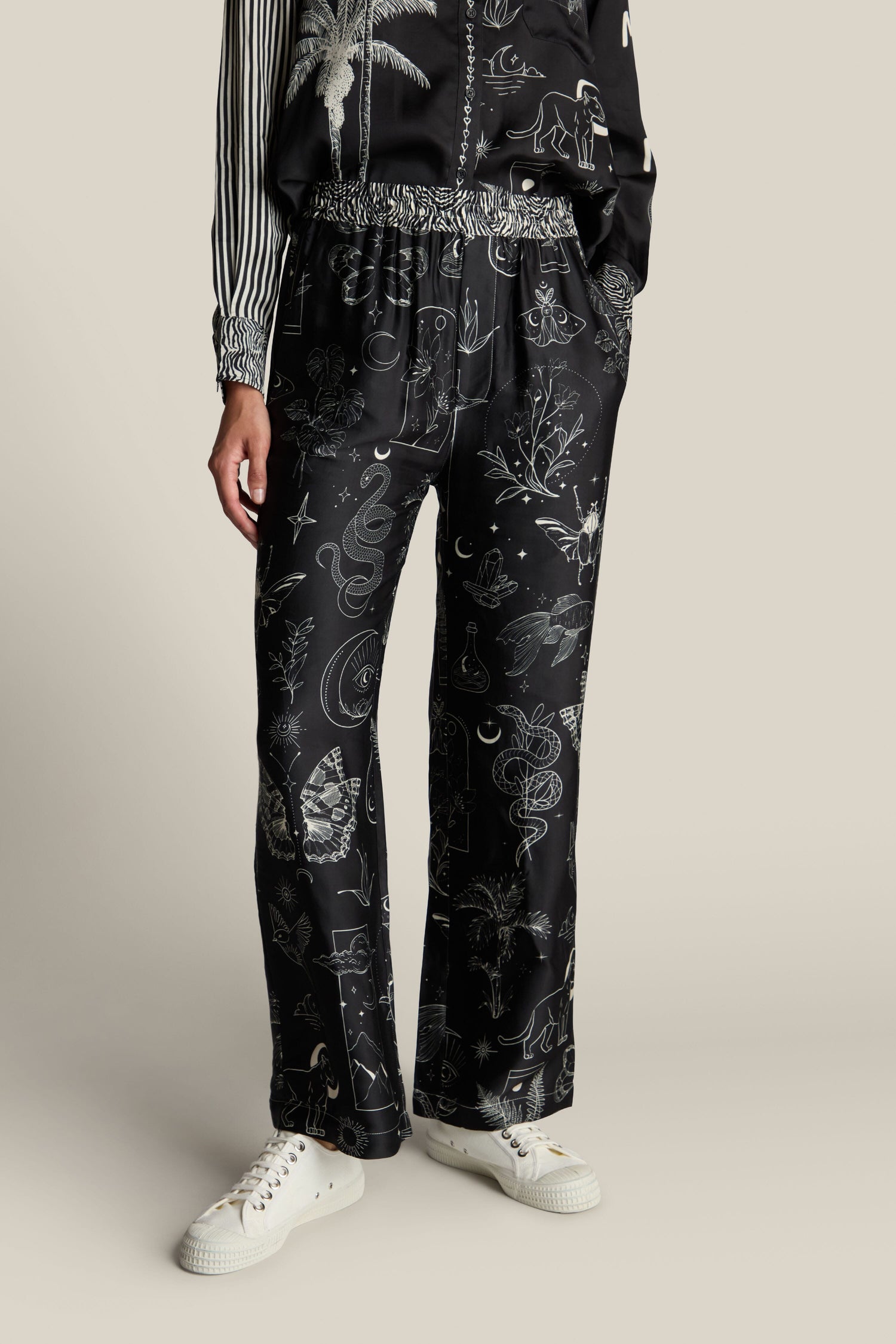 A person dressed in Black Magic Bailey Trouser, featuring a drawstring waist, paired with a mystical digital print top and white sneakers, stands against a plain background.