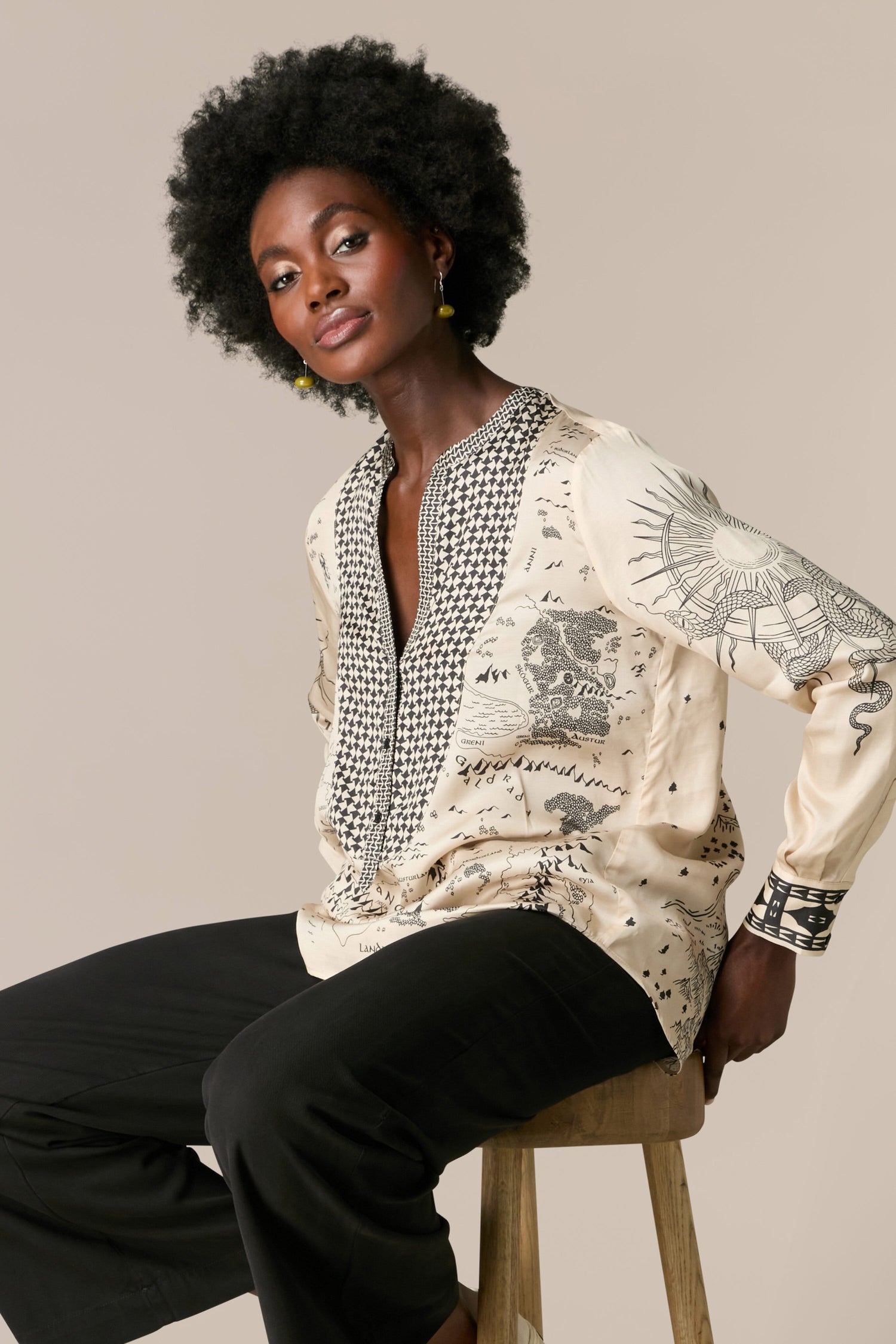 A black woman is sitting on a stool wearing a blouse in the Gabriella Bib Front Shirt.