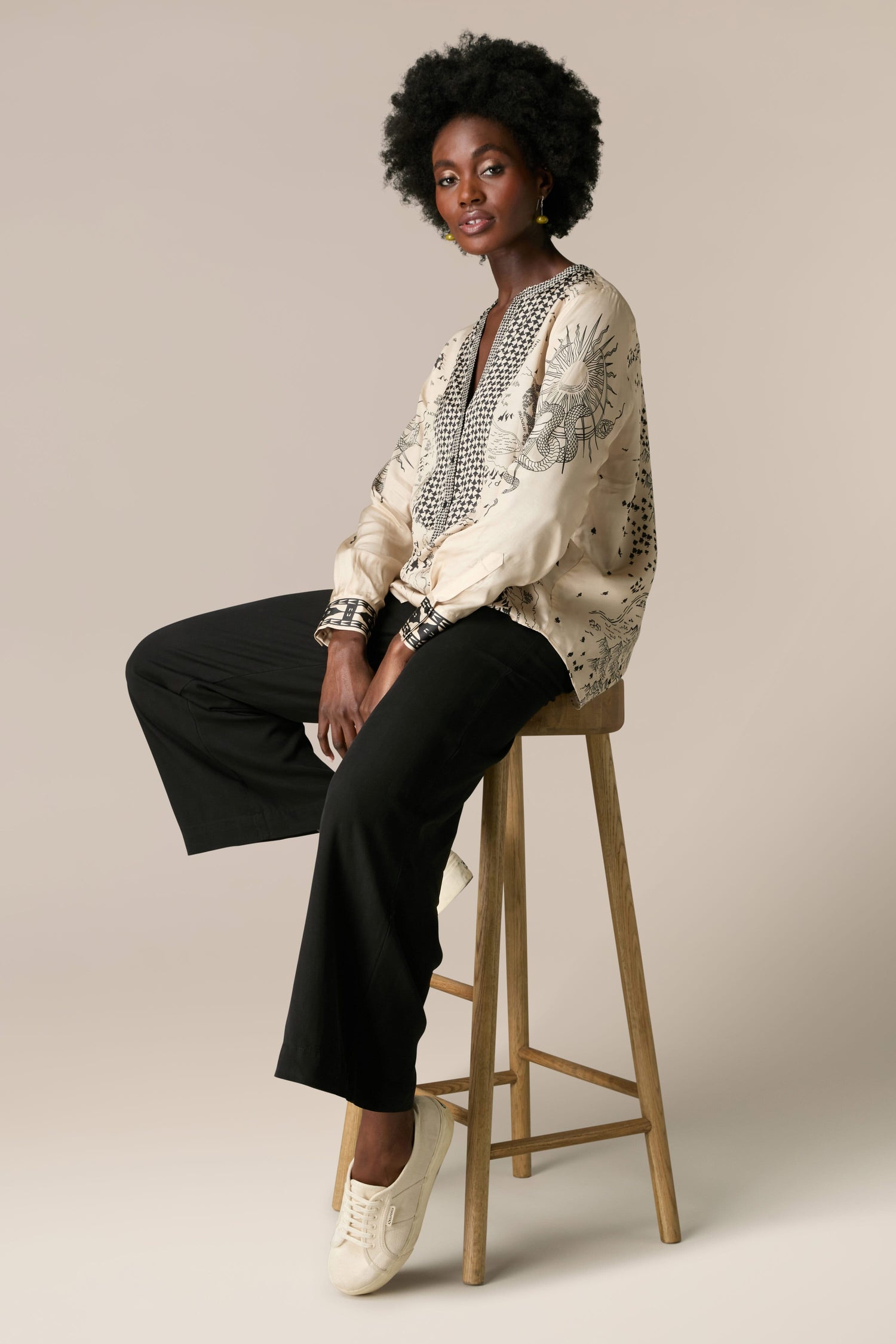 A black woman sitting on a stool wearing a blouse and pants, styled in the Gabriella Bib Front Shirt made of silky flowing fabric.