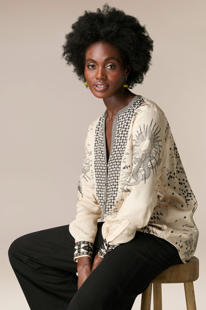 A woman with afro hair sitting on a stool, wearing the Gabriella Bib Front Shirt made of silky flowing fabric.