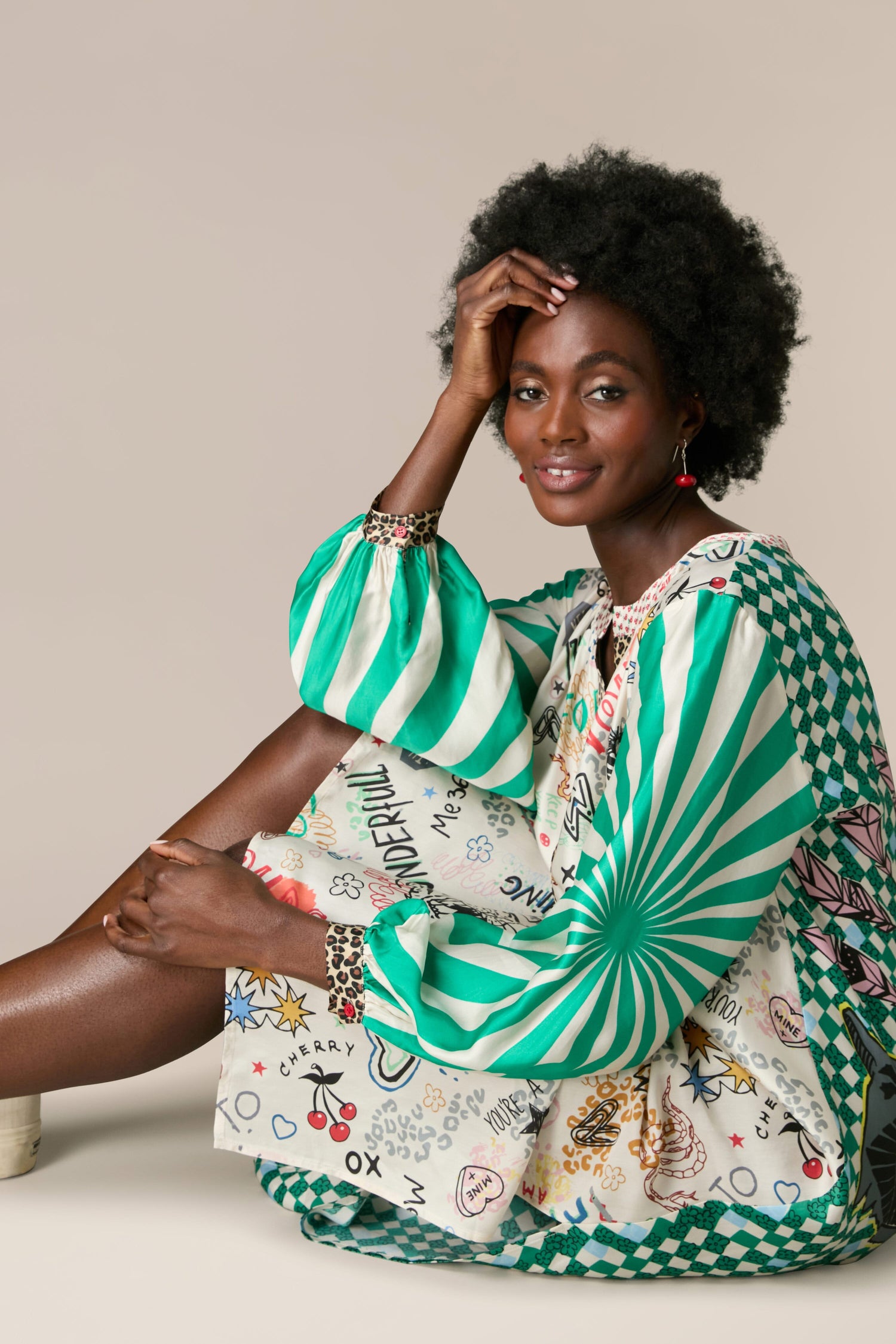 A black woman gracefully rests on the floor, dressed in a Hannah Deep V Neck Graffiti Dress. Her attire, made of silky flowing fabric, features a captivating ME369 graphic pattern in green and white.