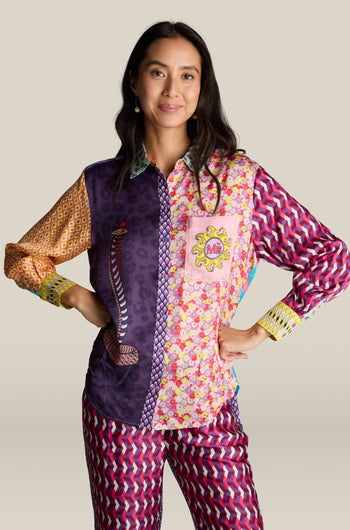 A woman wearing a vibrant Twisted Love Isabel Shirt, featuring intricate digital prints, paired with patterned pants, stands confidently with her hands on her hips against a beige background.