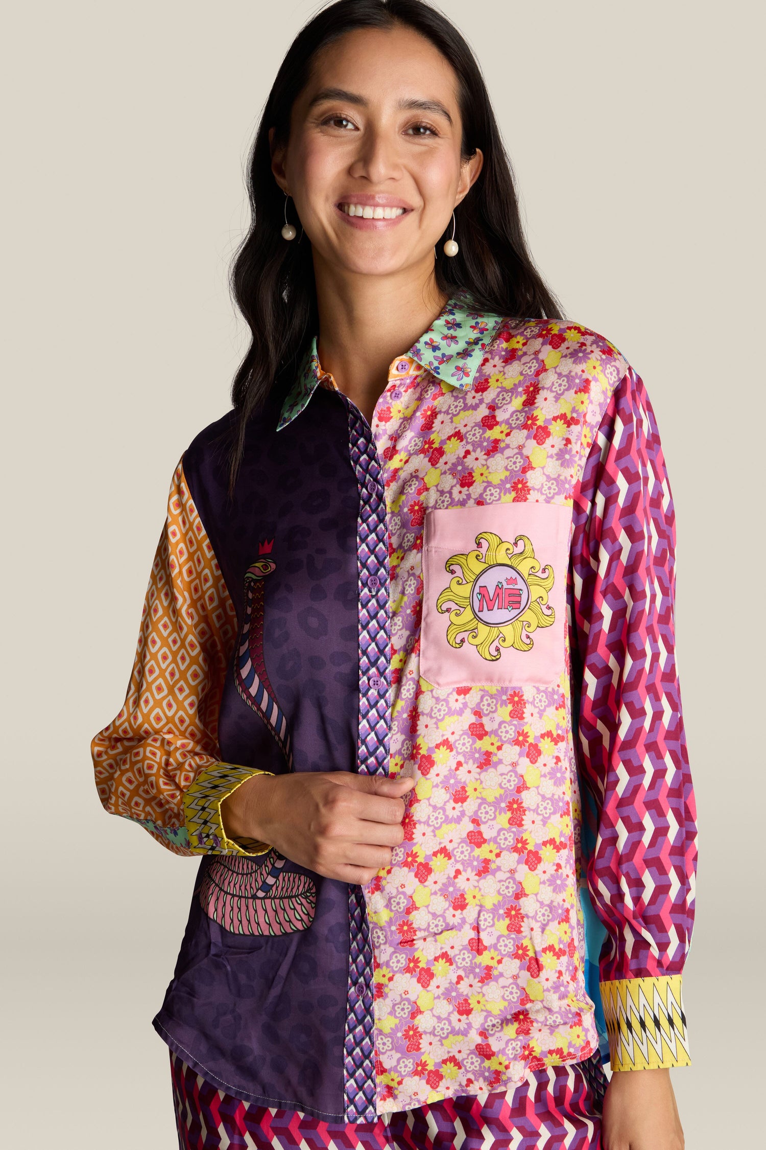 A woman smiles and wears a brightly patterned Twisted Love Isabel Shirt featuring designs that include a peacock, floral patterns, and snake illustrations, standing against a plain background.