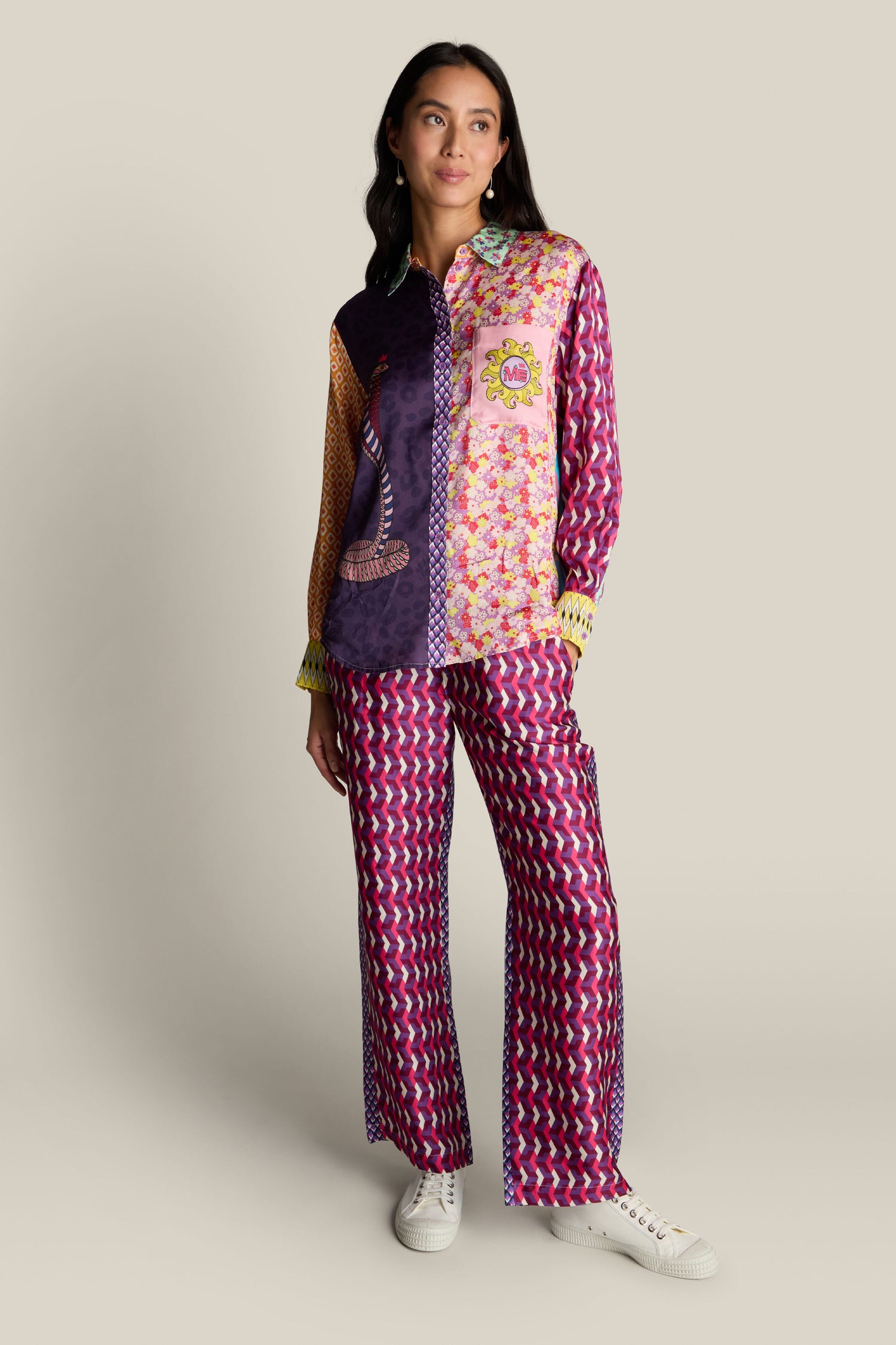 A woman stands against a plain background, wearing a colorful patterned outfit consisting of the Twisted Love Isabel Shirt with digital prints and matching wide-leg pants. With her hands in her pockets, she gazes off to the side, highlighting the intricate snake illustration on her shirt.