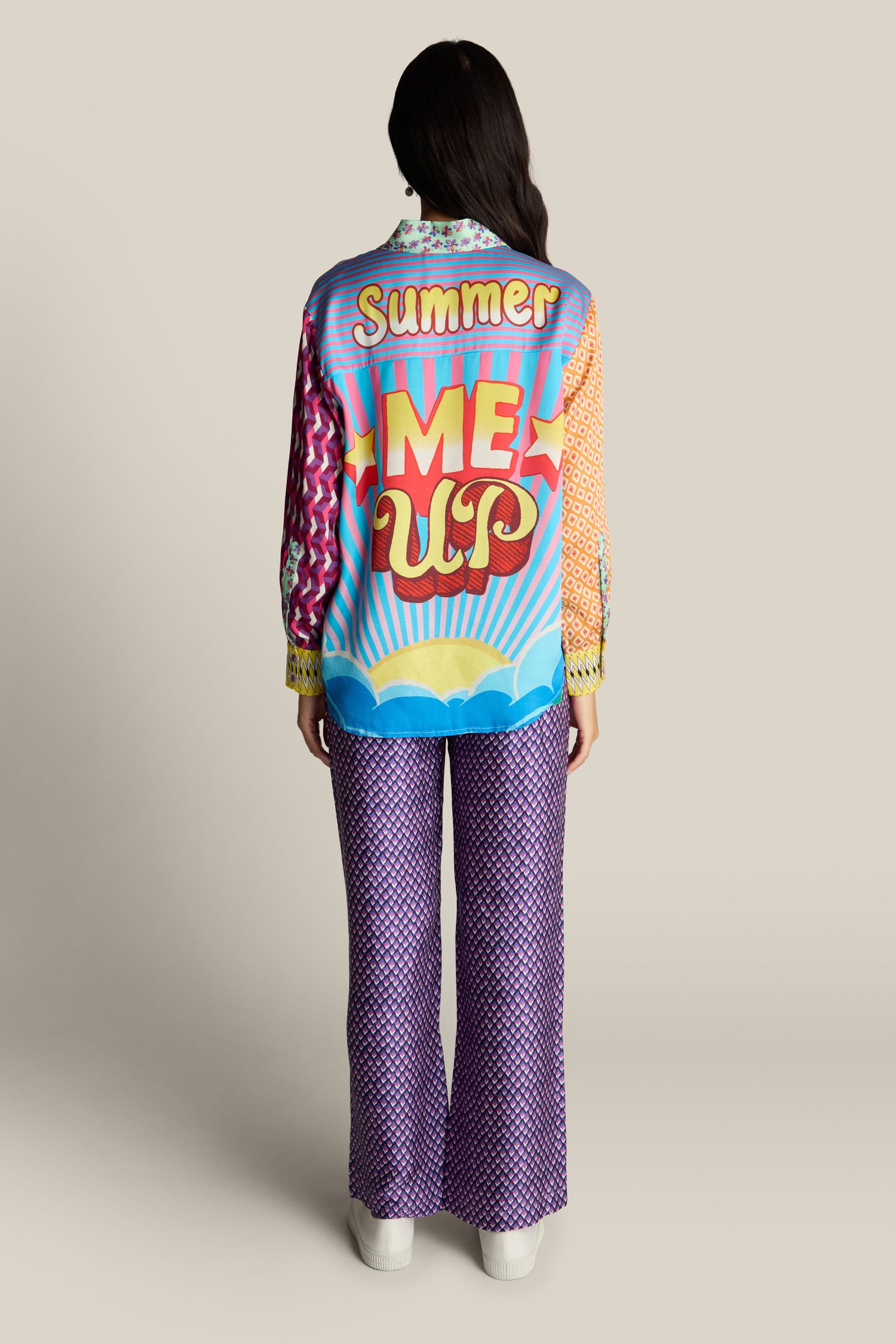 A person with long hair is facing away, wearing a colorful jacket with "Summer Me Up" text, patterned pants, and the distinctive style of a Twisted Love Isabel Shirt featuring digital prints.