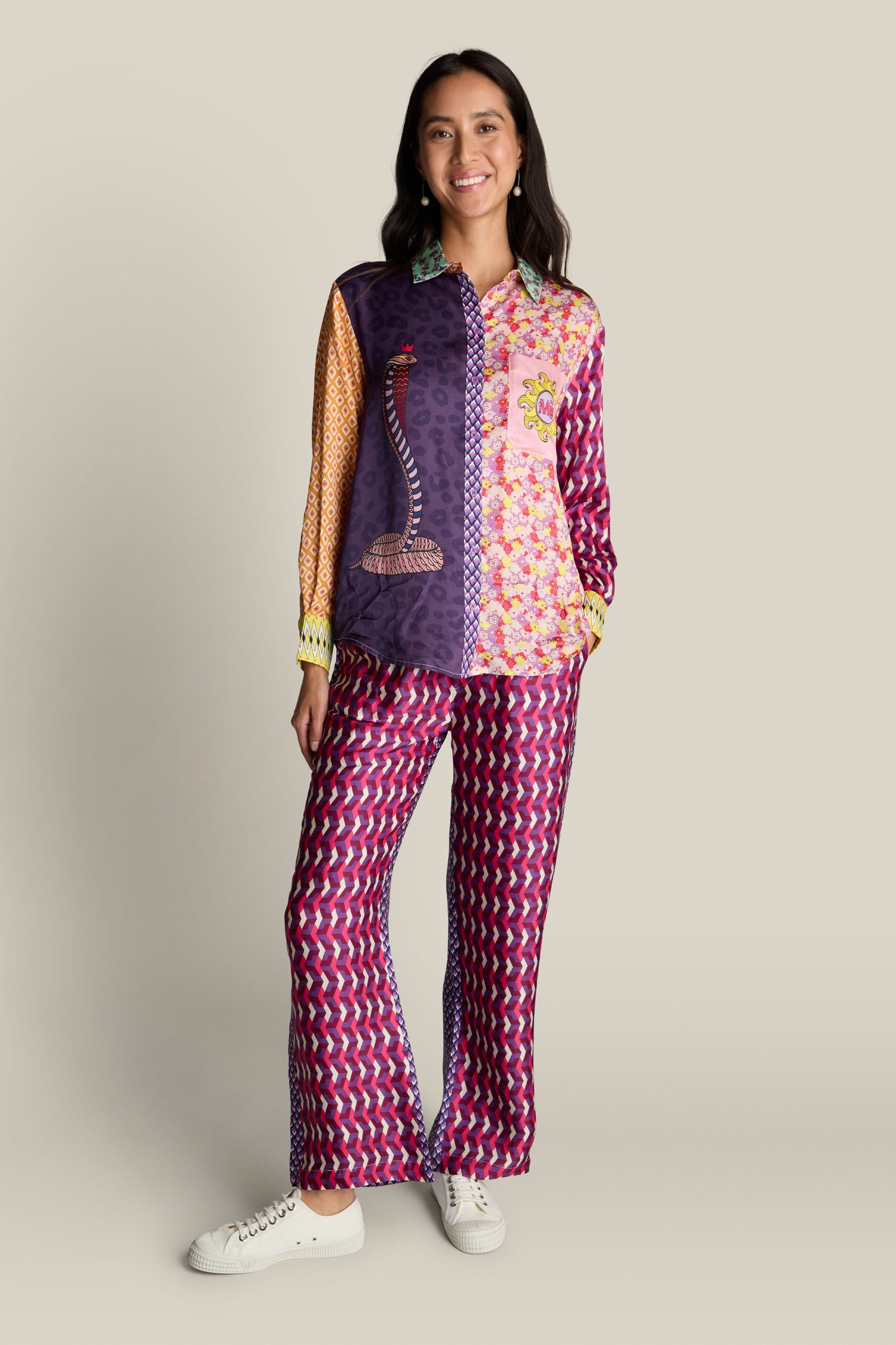 A person stands against a neutral background, wearing a vibrant outfit made up of the Twisted Love Isabel Shirt and matching pants adorned with vivid digital prints. They are smiling and have their hands in their pockets.