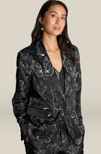 A person with long dark hair stands against a neutral background, wearing the Molly Printed Jacket featuring various white line drawings, elegantly highlighting its luxurious viscose and rayon fabric.