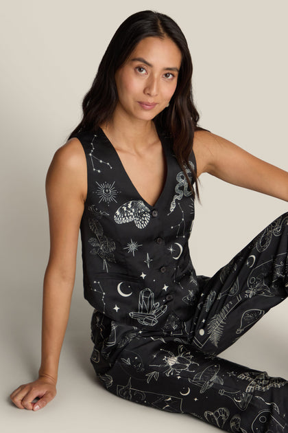 A woman with long dark hair sits on the floor, wearing a Cameron Vest, a stunning piece made from black fabric featuring intricate white celestial and mystical digital prints from ME369.